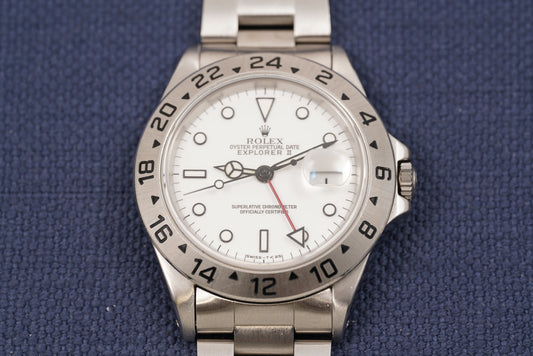 A Rolex Explorer II wristwatch with a stainless steel bracelet and bezel, featuring a white dial with black markings and a red seconds hand, displayed on a textured blue background.